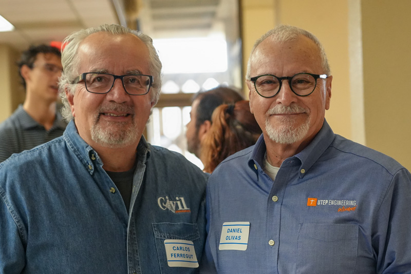 Two attendees wearing name badges pose together during a UTEP Engineering homecoming event, standing side by side in a gathering space while other guests mingle in the background.
